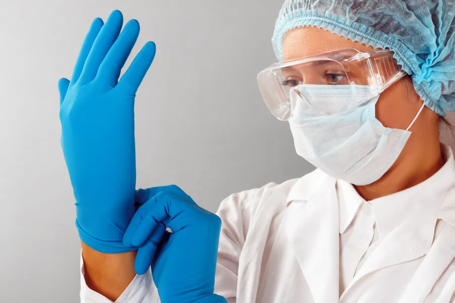 Laborfachkraft mit Haube, Maske und Schutzbrille zieht blaue Handschuhe an